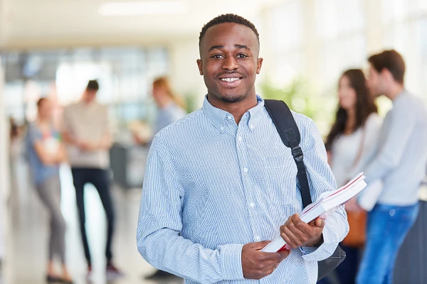 A smiling student with a backpack and books stands in a bright hallway with other students in the background.