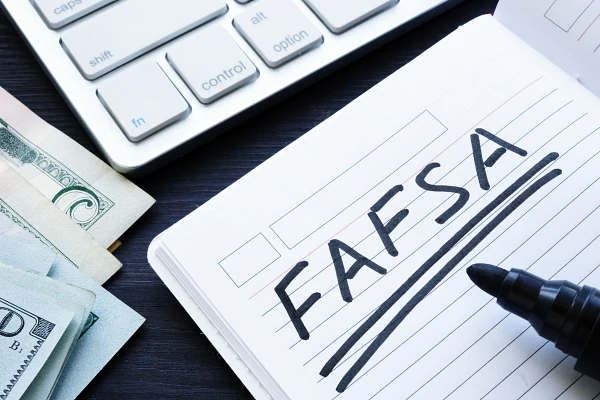 The image shows a notebook with "FAFSA" written on it, alongside a keyboard and a stack of cash.