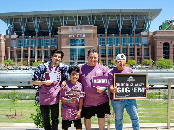 A family standing in front of Kyle Field holding props and signs for Family Weekend.