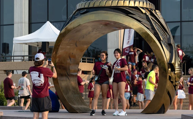Students gather around a large replica of a ring for a photo during an outdoor event.