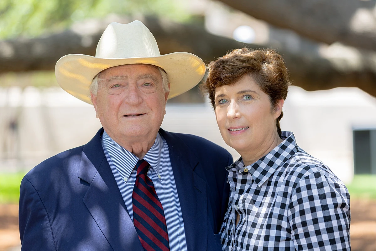 An older man in a cowboy hat and a suit stands beside a woman in a checkered shirt, both smiling outdoors.