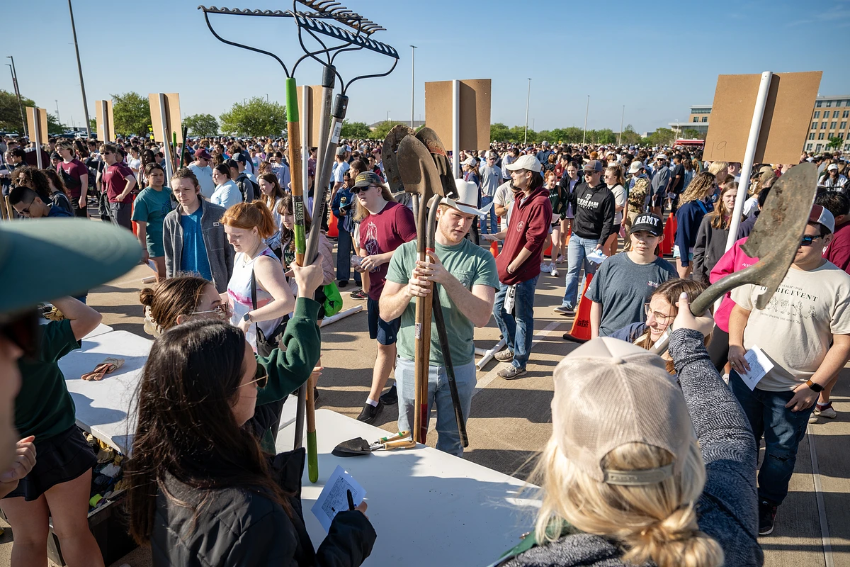 A large group of students gathered outdoors, some holding or distributing garden tools.