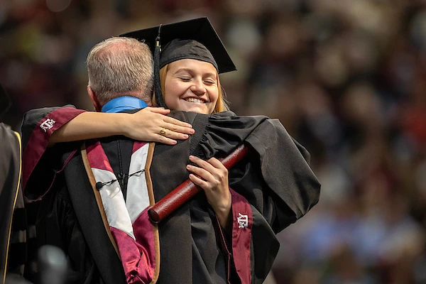 A graduate in a cap and gown is joyfully hugging another person during a ceremony.