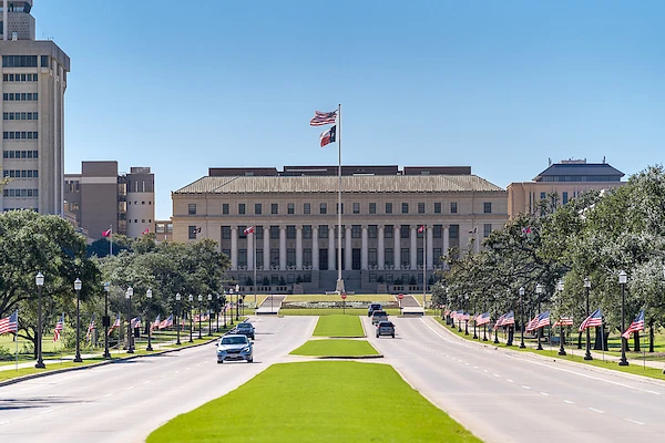 The image shows a symmetrical view of a historic building with columns, an American and Texas flag, and a tree-lined road leading up to it.