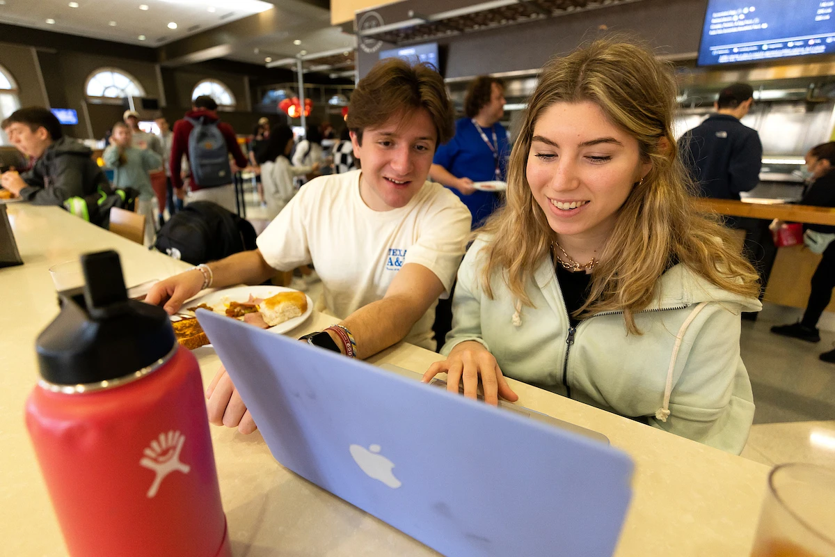 Two people smiling at a laptop in a busy cafeteria.