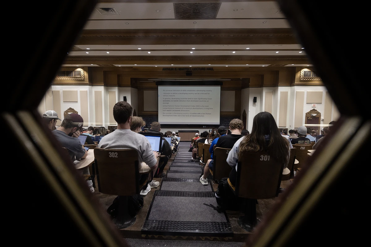 Students are seated in a large lecture hall, focusing on a presentation at the front.
