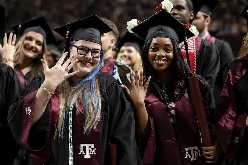 A group of graduates in caps and gowns celebrate while smiling and holding up rings.