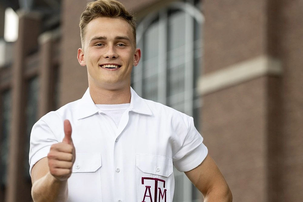 A person wearing a white shirt with an "A&M" emblem gives a thumbs-up in front of a brick building.