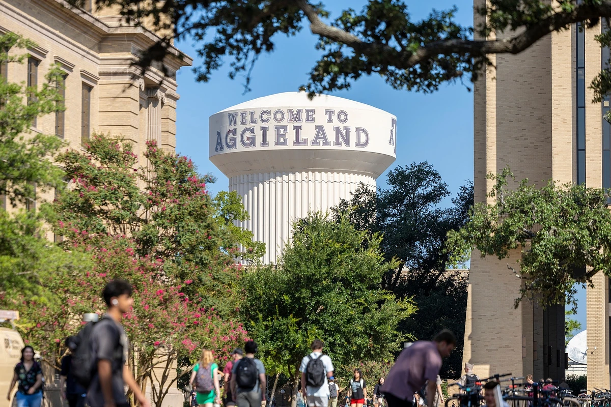 A water tower labeled "Welcome to Aggieland" is seen behind trees and people walking on a sunny day.