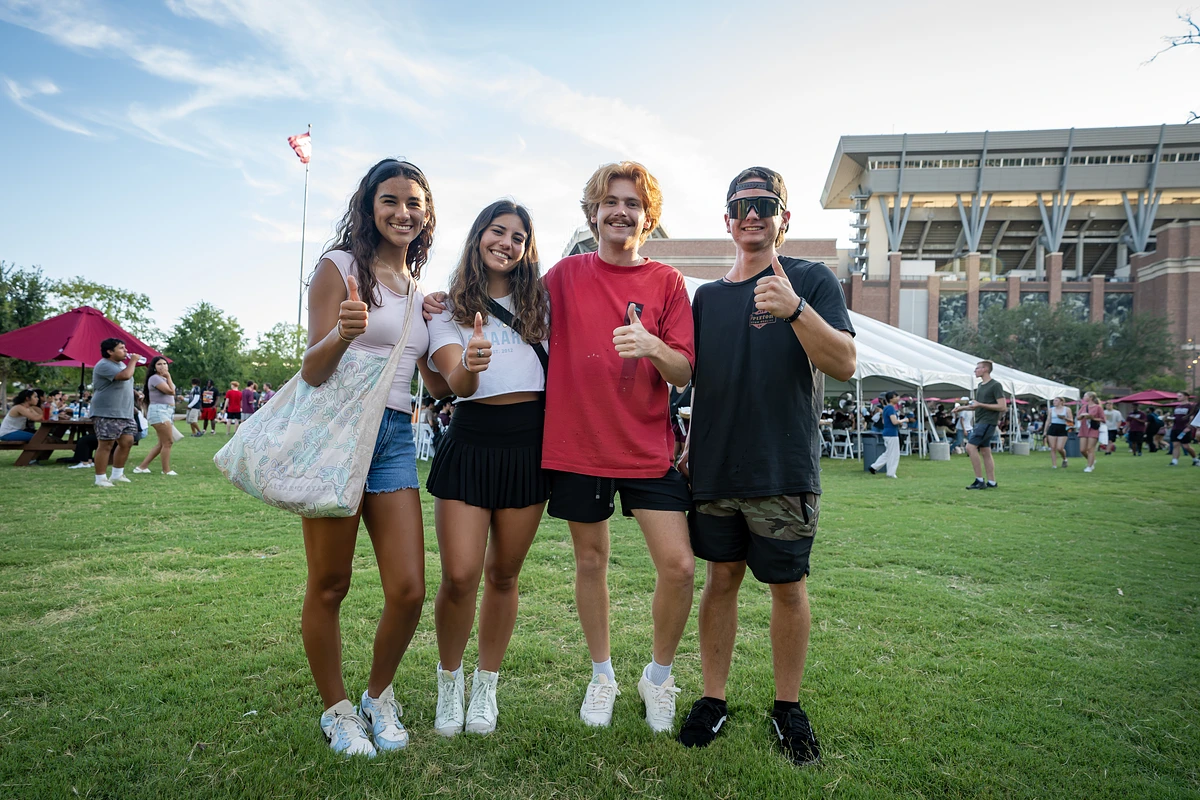 Four people are giving a gig'em (thumbs up) at an event in Aggie Park. 