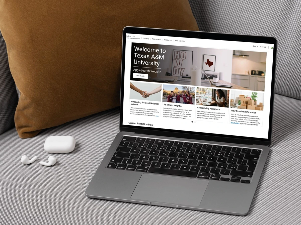 An open laptop sitting on a couch cushion with the AggieSearch home page open on the screen. A pair of wireless earbuds and a throw pillow sit in the background.