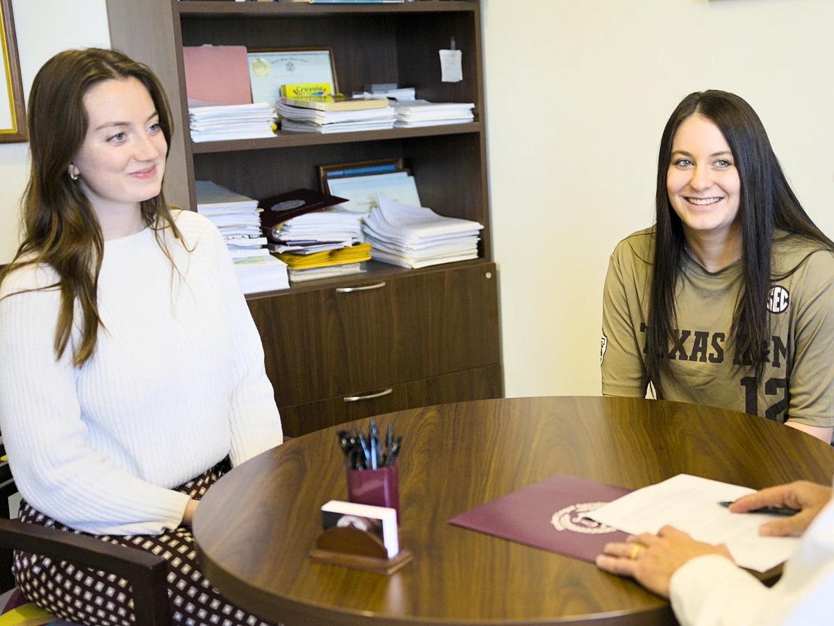 Two students sit at a small round table across from the Students’ Attorney holding a pen and documents, with a maroon Texas A&M folder on the table. Shelves filled with papers and folders are visible in the background.