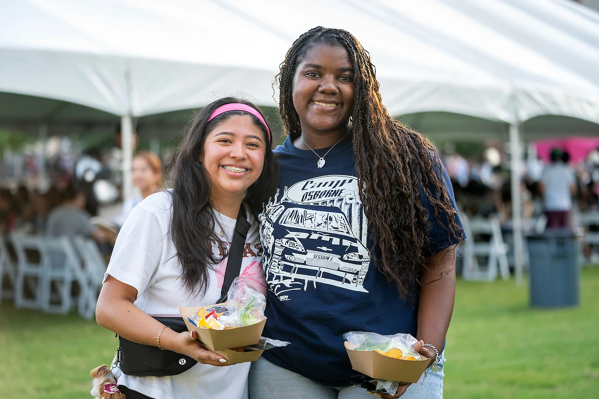 Two smiling people are holding trays of food in an outdoor setting with a large tent in the background.