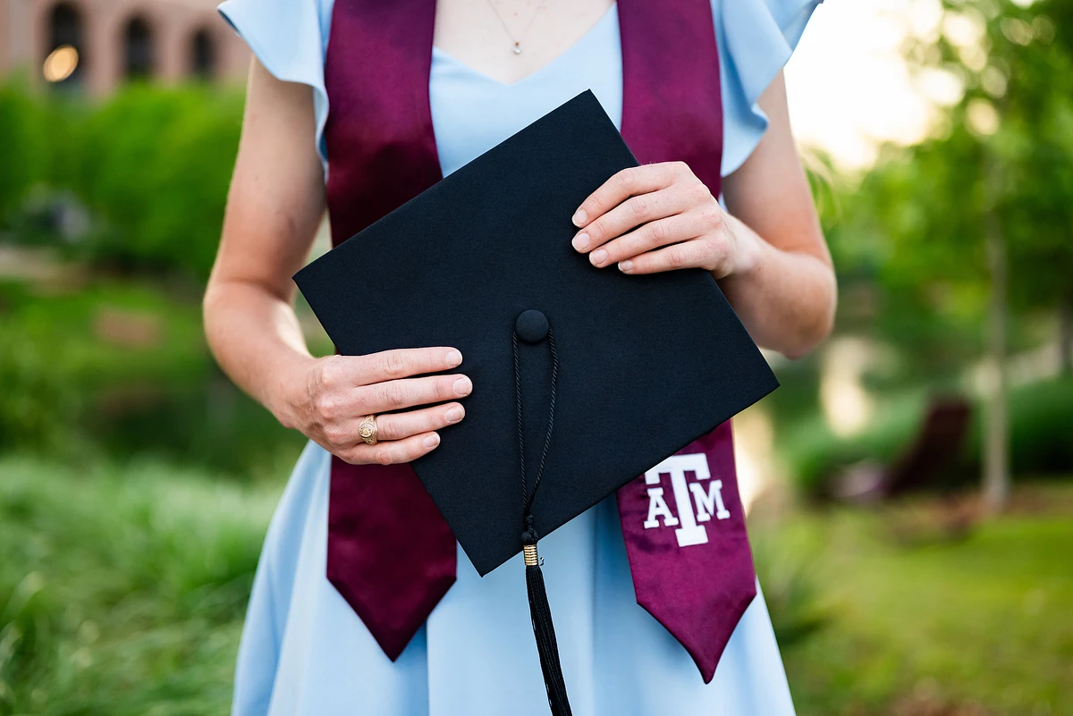 A person in a light blue dress and maroon stole, holding a black graduation cap in a green outdoor setting.